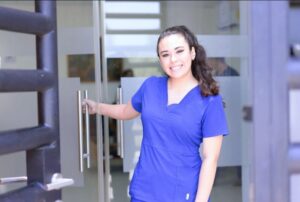 Smiling nurse in blue scrubs holding a door open, welcoming patients into a healthcare facility with a friendly and approachable demeanor.