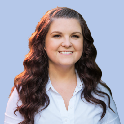 Portrait of a smiling woman with long wavy brown hair wearing a white blouse, set against a light blue background.